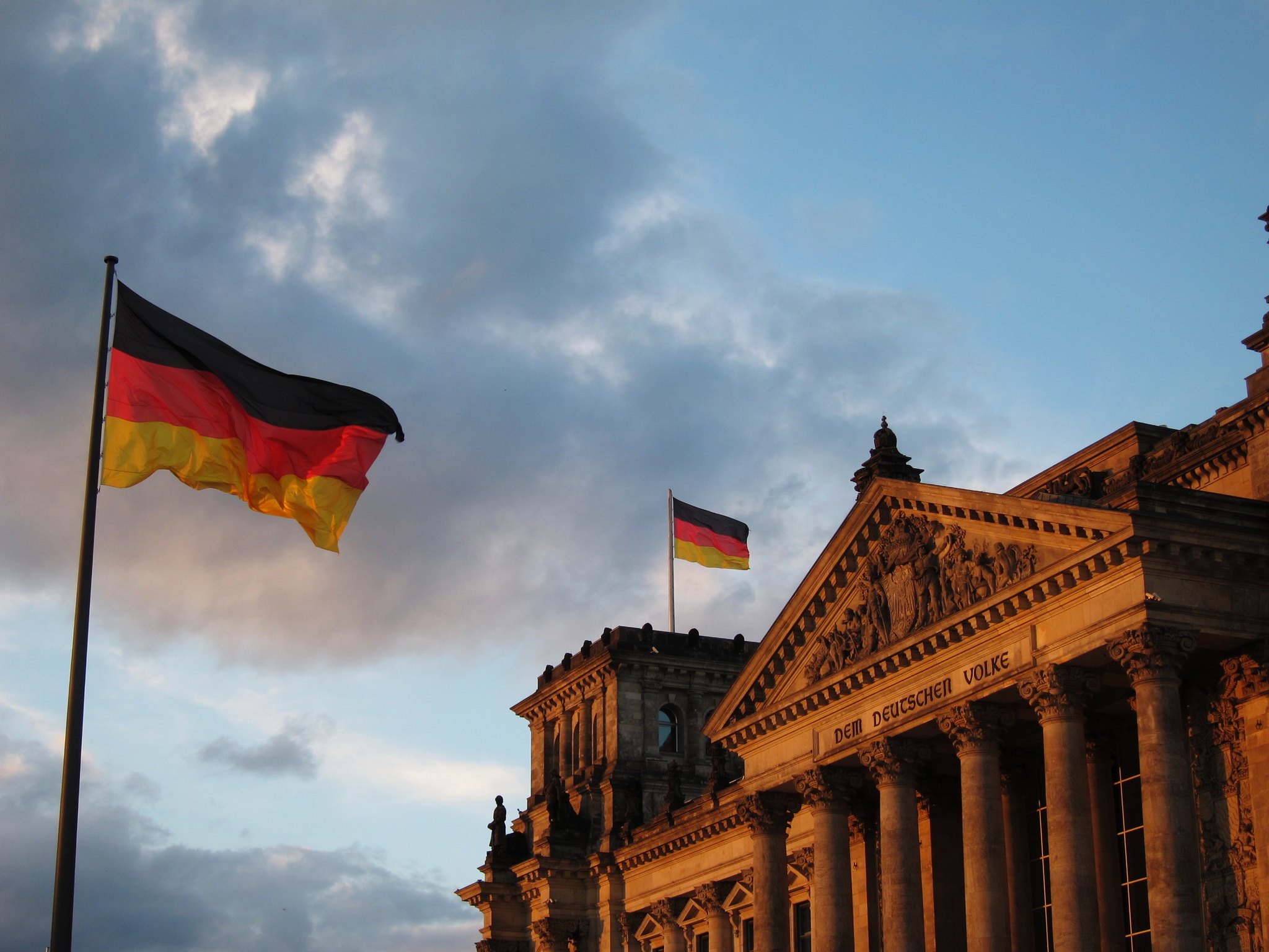 Na zdjęciu widać wielki budynek parlamentu Rzeszy w Berlinie, zawieszoną na nim niemiecką flagę i wielką niemiecką flagę obok po lewej stronie fotografii