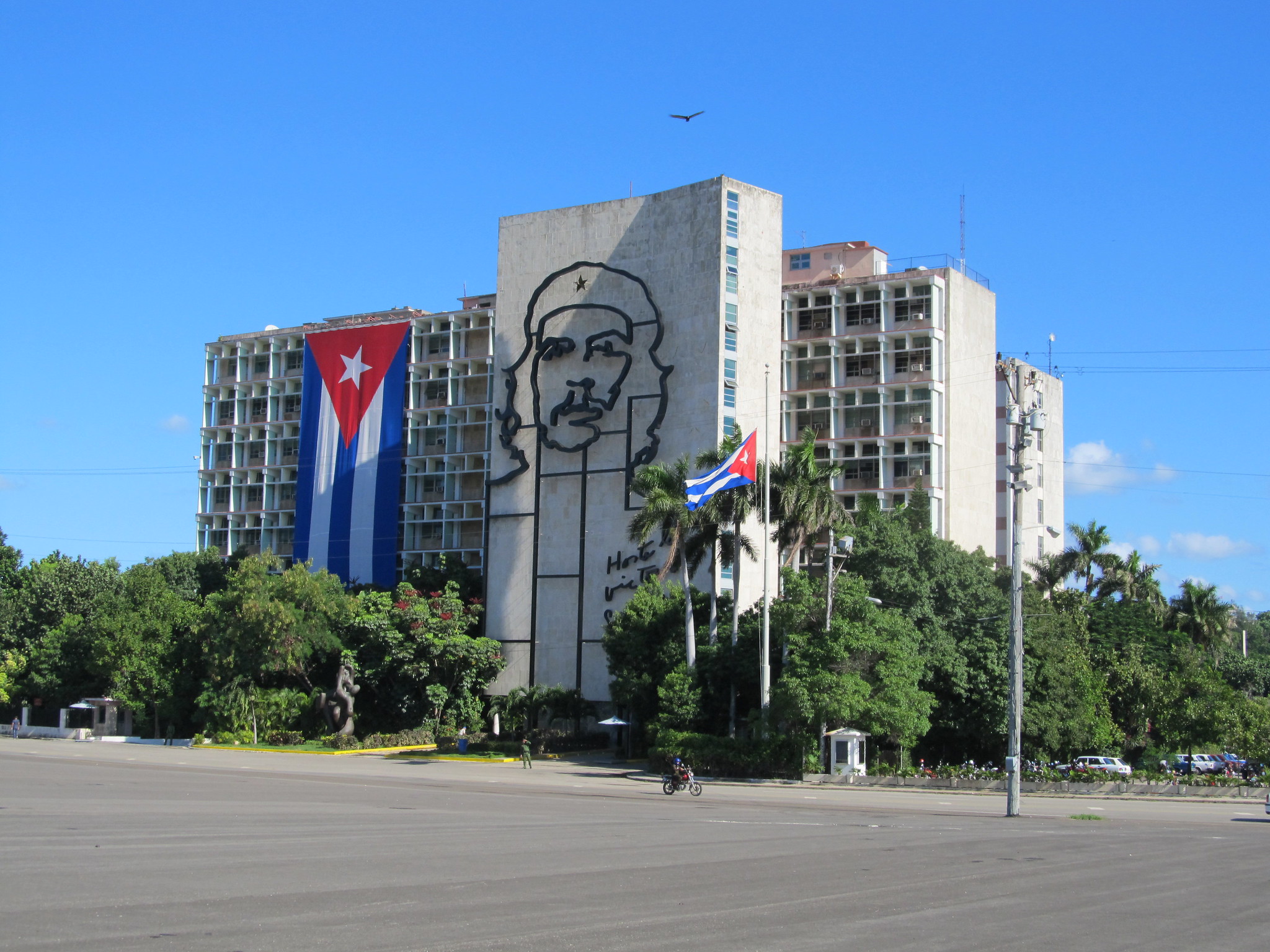 Zdjęcie przedstawia 3 budynki mieszkalne, które są położone wśród zielonych drzew. Na budynku wisi wielka flaga Kuby oraz wizerunek rewolucjonisty Che Guevary