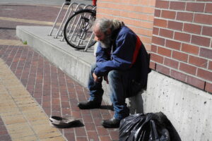 Na zdjęciu siedzi osoba bezdomna, przed nią leży czapka, do której zbiera pieniądze