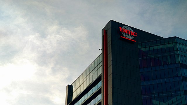 Fotografia przedstawia budynek, jednej z fabryk TSMC (tajwańskiego producenta półprzewodników), na budynku jest logo TSMC. W tle widać niebo.