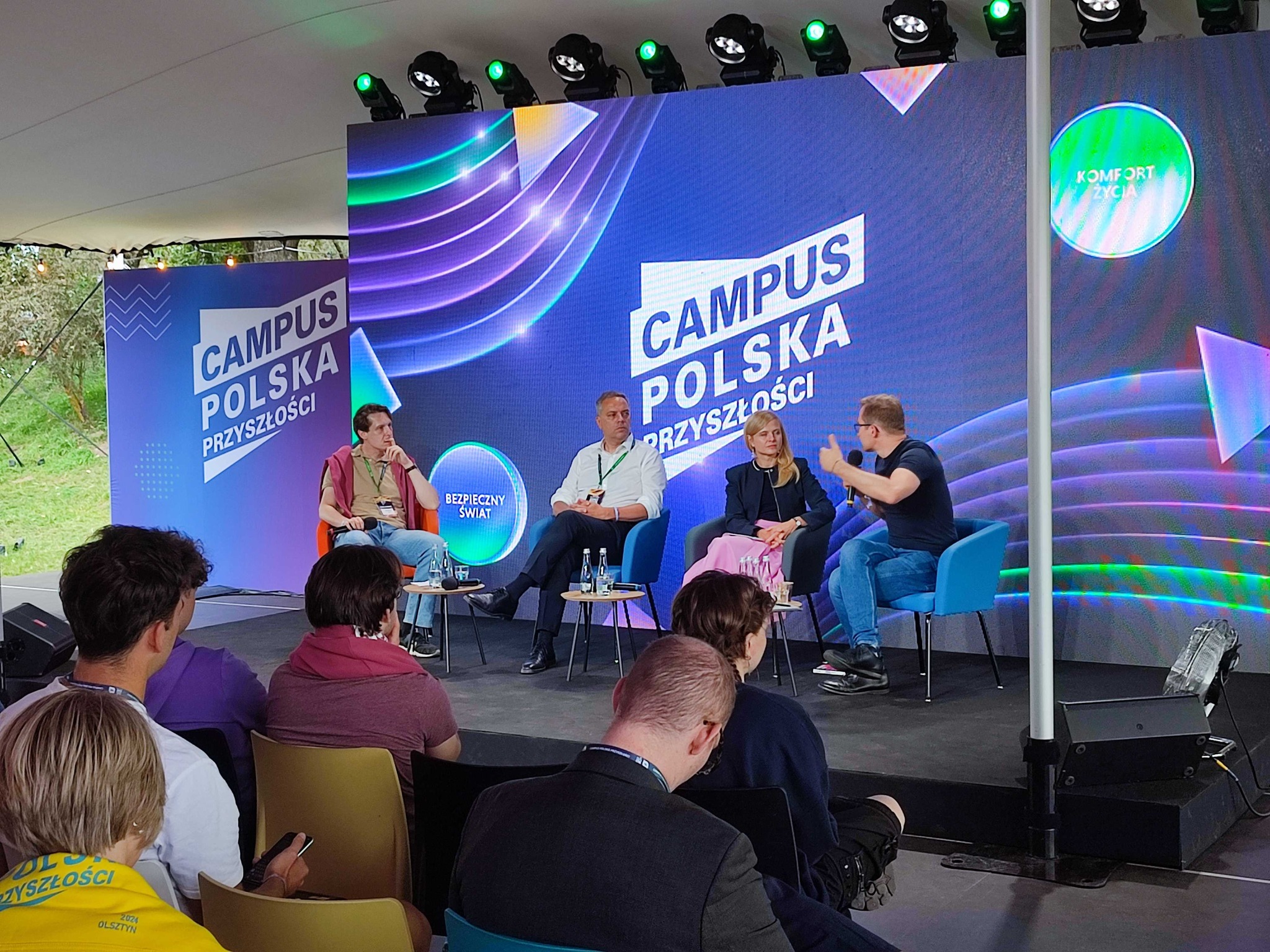 Dyskusja panelowa na wydarzeniu Campus Polska Przyszłości z czterema uczestnikami siedzącymi na scenie. Scena jest udekorowana dużym, kolorowym tłem z nazwą wydarzenia. Na pierwszym planie widoczna jest publiczność, która uważnie słucha prelegentów