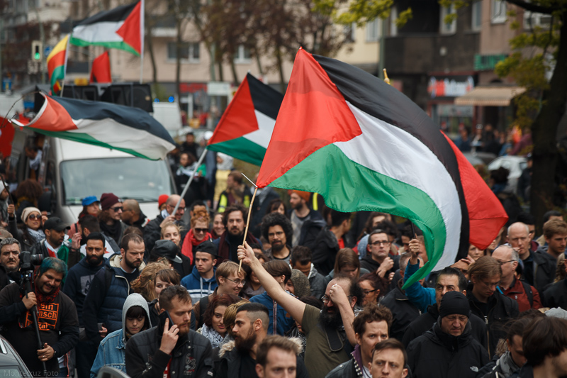 Duży tłum ludzi bierze udział w demonstracji, wiele osób trzyma i macha palestyńskimi flagami. Flagi mają kultowe kolory: czerwony, czarny, biały i zielony. Scena wydaje się rozgrywać na obszarze miejskim, w tle widać budynki i drzewa. Atmosfera wydaje się intensywna, a różnorodna grupa uczestników wyraża solidarność.