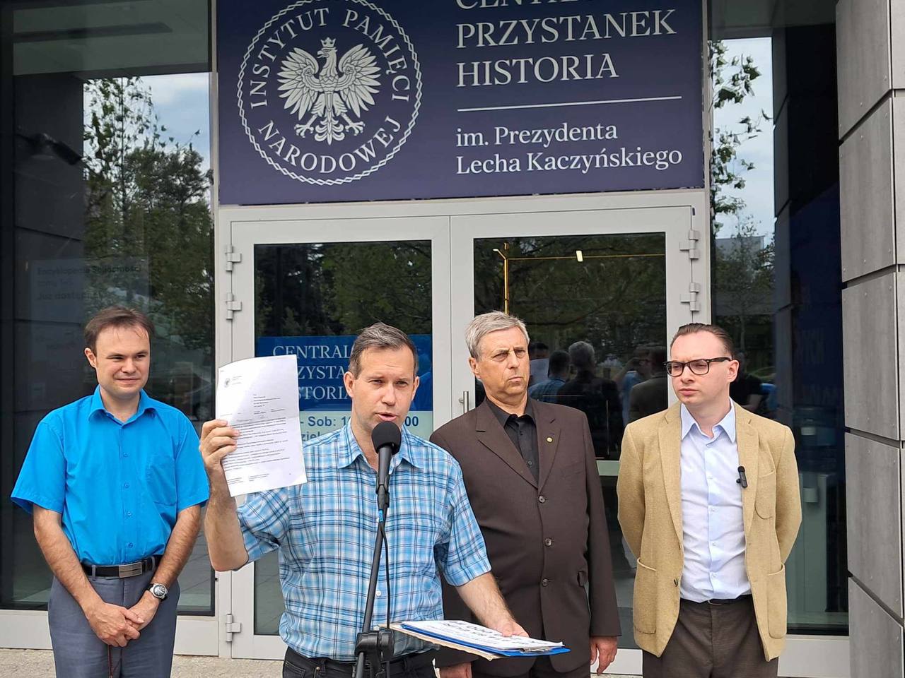 Czterech mężczyzn stoi przed wejściem do budynku Centralnego Przystanku Historia im. Prezydenta Lecha Kaczyńskiego. Jeden z nich, trzymający mikrofon, unosi dokument w ręce, podczas gdy pozostałe osoby patrzą na niego. W tle widoczny jest szyld Instytutu Pamięci Narodowej oraz nazwa placówki.