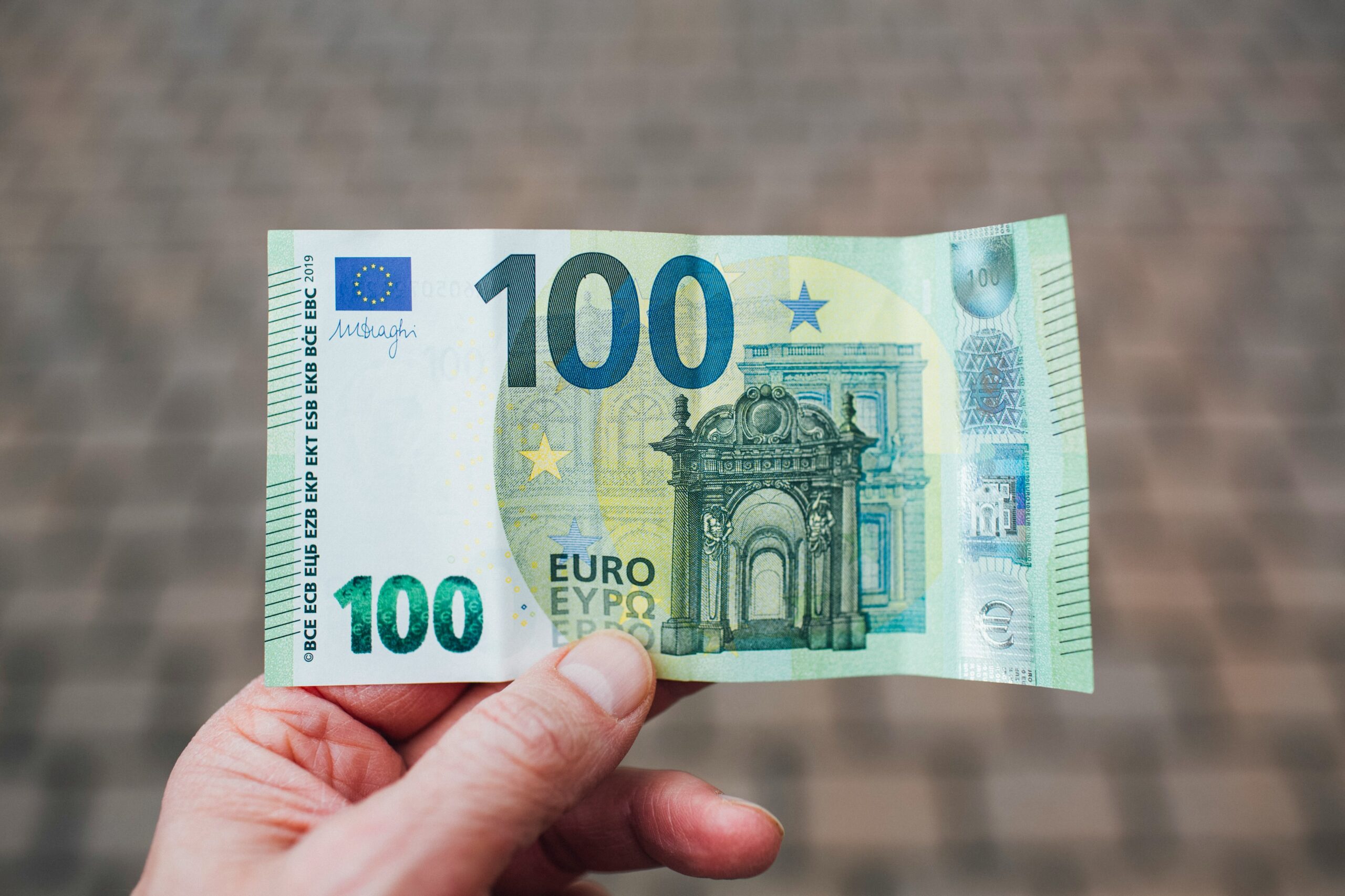 Osoba trzymająca w palcach banknot o nominale 100 euro. Na przedniej stronie banknotu widnieją motywy architektoniczne, w tym łuk oraz duża liczba "100" w zielonym kolorze. W lewym górnym rogu znajduje się podpis prezesa Europejskiego Banku Centralnego oraz flaga Unii Europejskiej. Tło jest rozmyte.